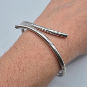以下が含まれることがあります： 手首に巻き付くように重なり合う、滑らかで丸みを帯びた2本のバンドを備えたバイパスデザインの、磨き上げられたシルバーカラーのメタルカフブレスレット。このブレスレットは光沢のある金属仕上げで、シンプルなオープンエンド構造になっています。