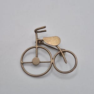 以下が含まれることがあります： 自転車の形をしたブロンズ調の金属製ブローチ。中央にハブがある大きな前輪、小さな後輪、湾曲したハンドルバー、そして平らなサドルが特徴です。金属はマットで少し使い古されたような質感に仕上げられています。