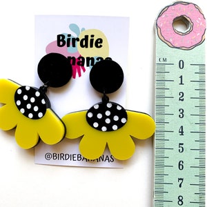 May include: A pair of yellow flower-shaped earrings with black polka dot accents and black round tops. The earrings are approximately 6 cm long. The earrings are displayed on a white background with a ruler for size reference. The text "Birdie nanas" and "@BIRDIEBANANAS" are visible on the card.