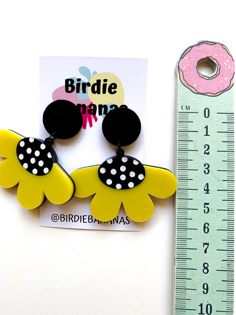 May include: A pair of yellow flower-shaped earrings with black polka dot accents and black round tops. The earrings are approximately 6 cm long. The earrings are displayed on a white background with a ruler for size reference. The text "Birdie nanas" and "@BIRDIEBANANAS" are visible on the card.