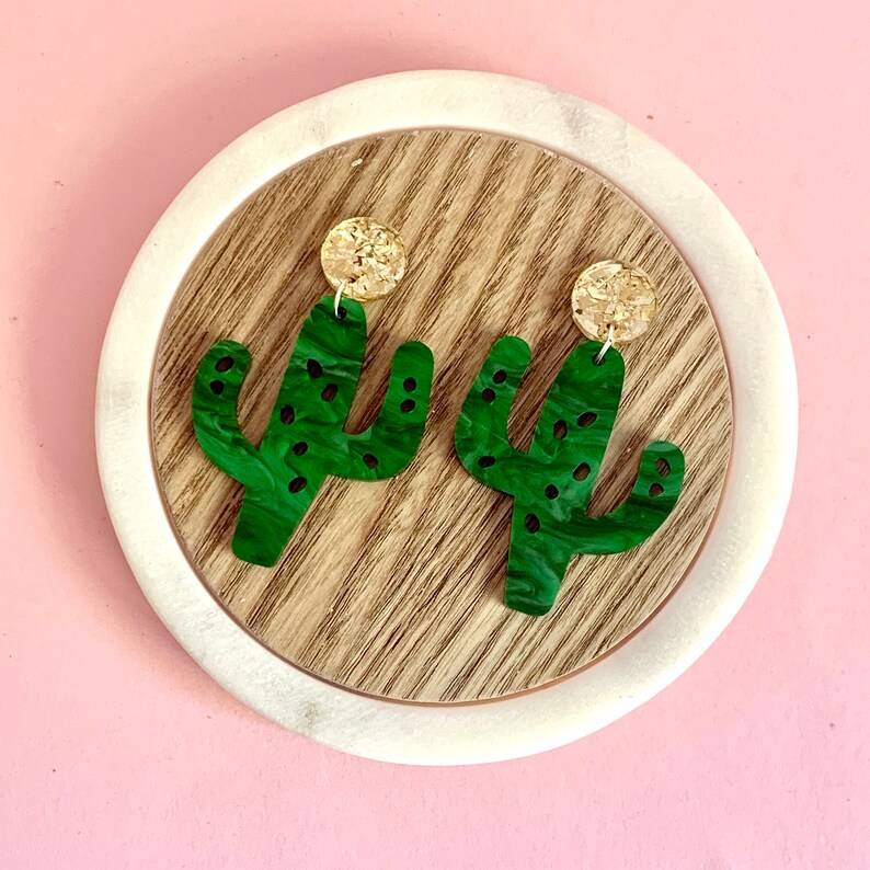 May include: A pair of green cactus-shaped earrings with cork accents. The earrings are on a wooden surface.