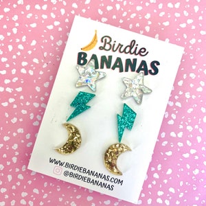 Peut inclure: Un ensemble de six boucles d'oreilles clous à paillettes avec un motif étoile, éclair et croissant de lune. Les boucles d'oreilles sont présentées sur une carte blanche avec le texte "Birdie Bananas" et des informations sur le site Web.