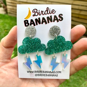K&ouml;nnte beinhalten: Ein Paar gr&uuml;ne Glitzer-Wolkenohrringe mit regenbogenfarbenen Blitzen als Anh&auml;nger. Die Ohrringe befinden sich auf einer wei&szlig;en Karte mit dem Text "Birdie Bananas" und einer Bananengrafik.