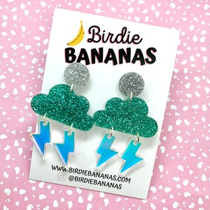 K&ouml;nnte beinhalten: Ein Paar gr&uuml;ne Glitzer-Wolkenohrringe mit regenbogenfarbenen Blitzen als Anh&auml;nger. Die Ohrringe befinden sich auf einer wei&szlig;en Karte mit dem Text "Birdie Bananas" und einer Website-Adresse.