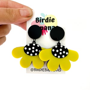 May include: A pair of yellow flower-shaped earrings with black and white polka dot accents. The earrings are hanging from black circles. The earrings are on a white card with the text "Birdie Banana" and "@birdiebananas".