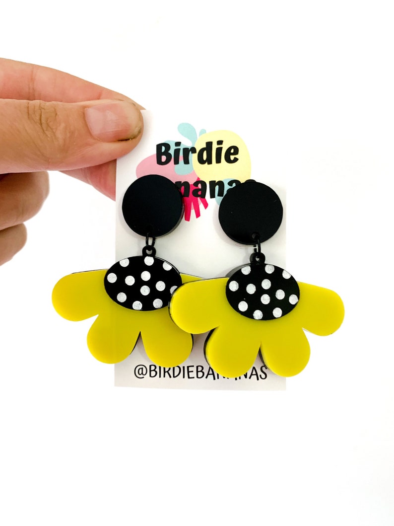 May include: A pair of yellow flower-shaped earrings with black and white polka dot accents. The earrings are hanging from black circles. The earrings are on a white card with the text "Birdie Banana" and "@birdiebananas".