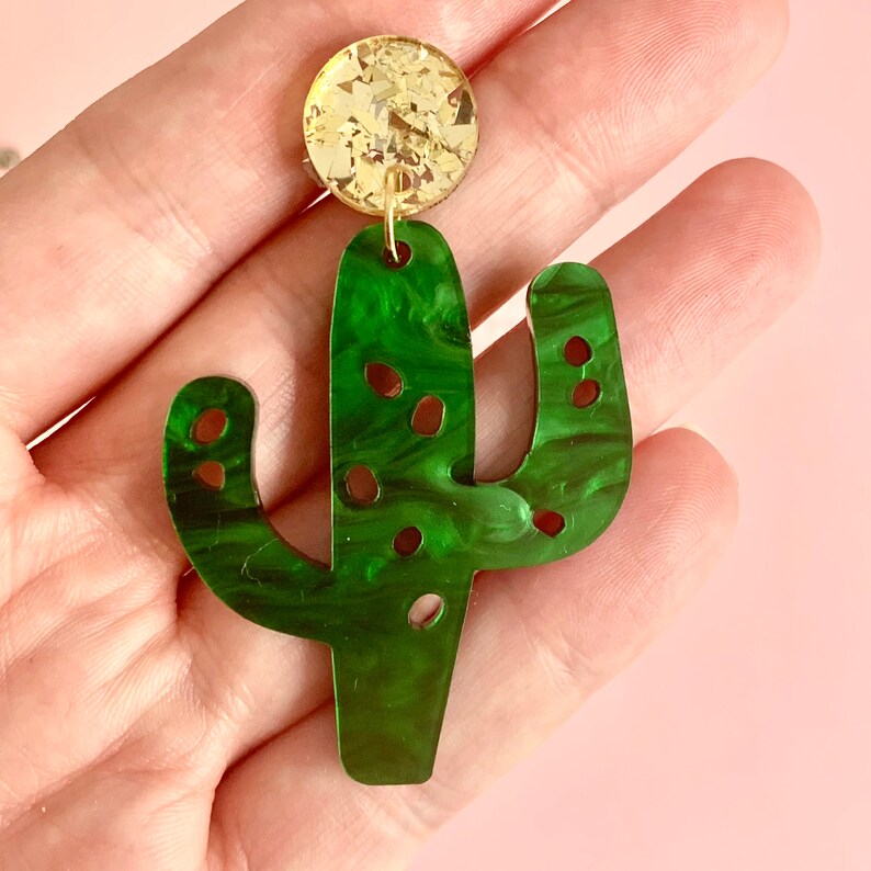 May include: Green cactus-shaped earrings with a gold glitter accent. The earrings are made of acrylic and have a marbled effect.