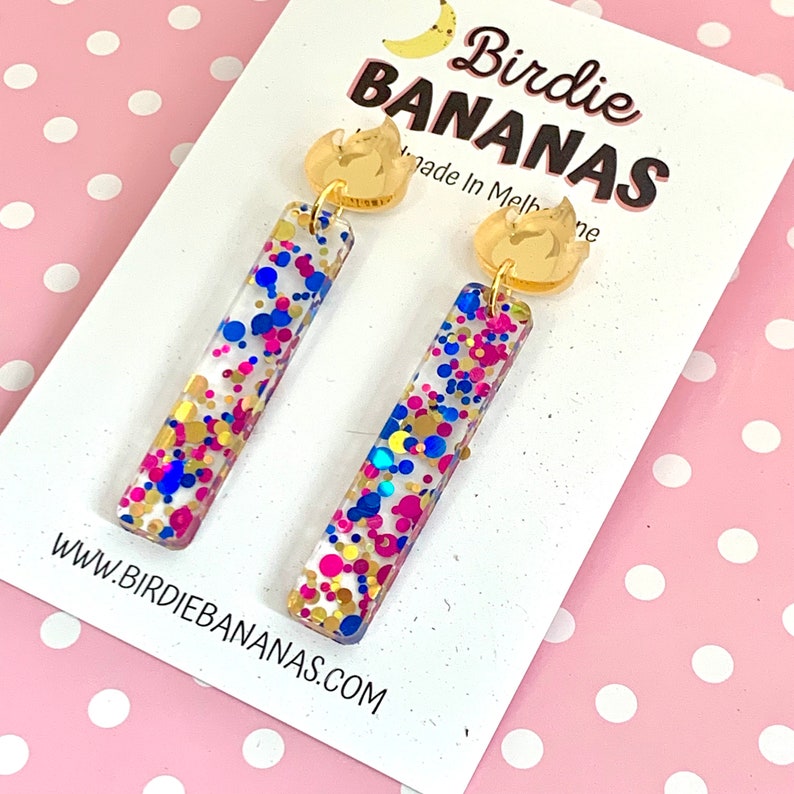 May include: A pair of gold and clear acrylic earrings with a confetti design. The earrings are shaped like a rectangle and have a gold flame charm at the top. The confetti is in shades of blue, pink, and gold. The earrings are displayed on a white card with the text "Birdie Bananas" and "Made in Melbourne".
