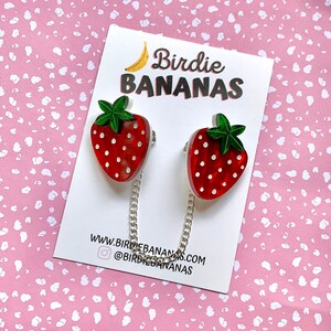 Peut inclure: Deux broches en forme de fraise rouge et blanche avec des feuilles vertes et des pois blancs reli&eacute;es par une cha&icirc;ne argent&eacute;e. Les broches sont sur une carte blanche avec le texte "Birdie BANANAS" et "www.birdiebananas.com @birdiebananas".