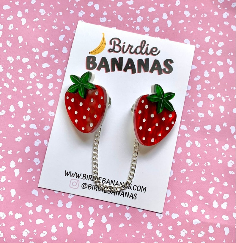 Peut inclure: Deux broches en forme de fraise rouge et blanche avec des feuilles vertes et des pois blancs reli&eacute;es par une cha&icirc;ne argent&eacute;e. Les broches sont sur une carte blanche avec le texte "Birdie BANANAS" et "www.birdiebananas.com @birdiebananas".