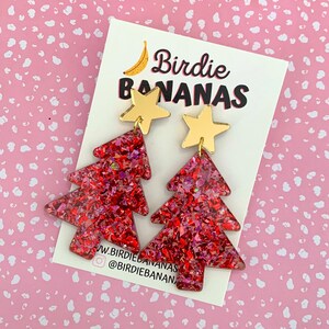 May include: A pair of Christmas tree earrings with a red glitter design and gold star toppers. The earrings are displayed on a white card with the text "Birdie Bananas". The background is pink with white polka dots.
