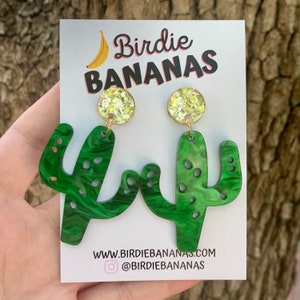 May include: Green cactus-shaped dangle earrings with gold glitter accents. The earrings are on a white card with the text "Birdie BANANAS" and a banana graphic. The card also includes the website address "www.birdiebananas.com" and the social media handle "@birdiebananas".