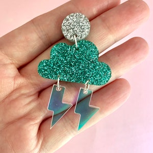 K&ouml;nnte beinhalten: Gr&uuml;n glitzernde Ohrringe in Wolkenform mit schimmernden Blitzen, die an der Unterseite h&auml;ngen. Die Ohrringe sind oben silberfarben.