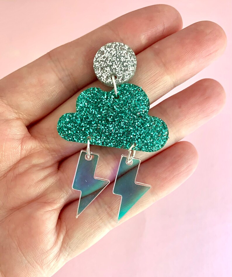 K&ouml;nnte beinhalten: Gr&uuml;n glitzernde Ohrringe in Wolkenform mit schimmernden Blitzen, die an der Unterseite h&auml;ngen. Die Ohrringe sind oben silberfarben.