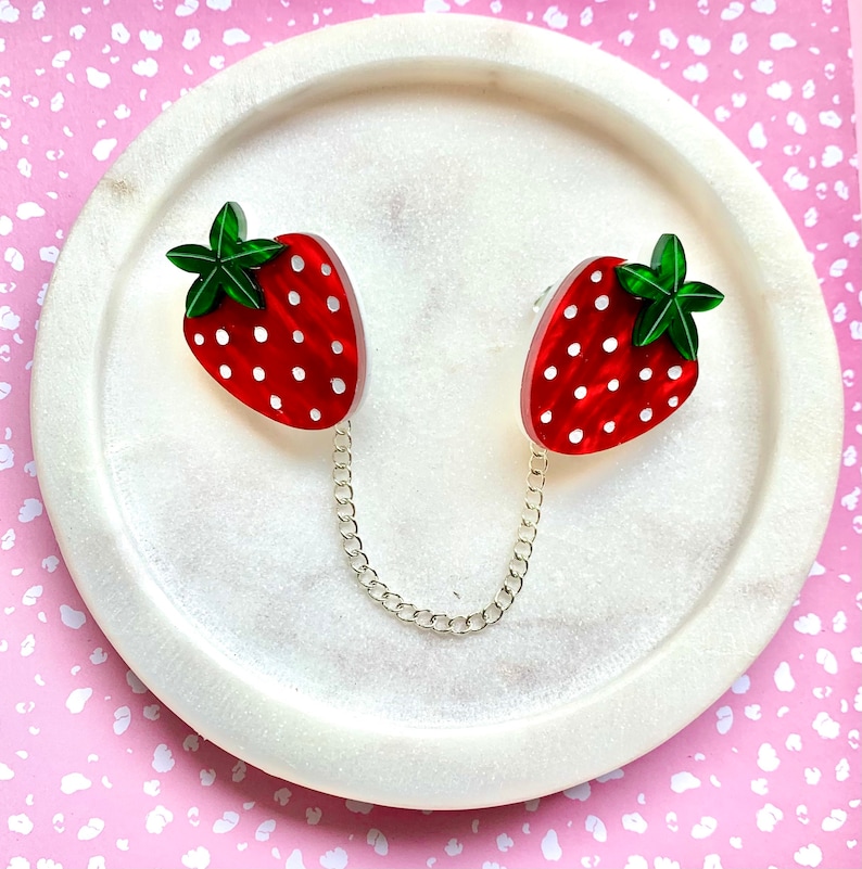 Peut inclure: Deux boucles d'oreilles en forme de fraise rouge et blanche avec des feuilles vertes reli&eacute;es par une cha&icirc;ne argent&eacute;e. Les boucles d'oreilles sont sur une surface en marbre blanc.