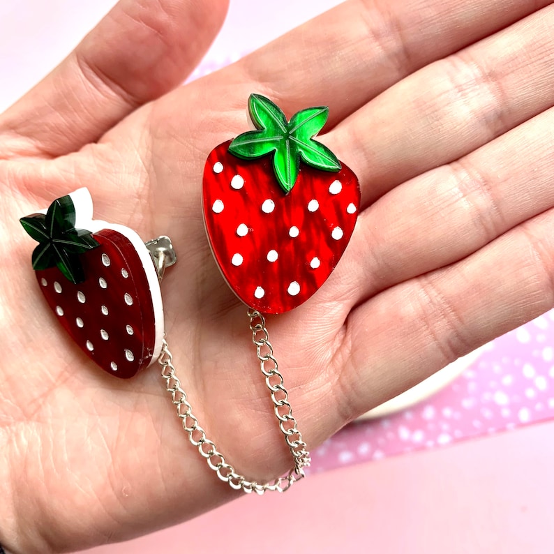 Peut inclure: Une paire de broches en forme de fraises rouges et blanches reli&eacute;es par une cha&icirc;ne argent&eacute;e. Les fraises ont des feuilles vertes et des points blancs.