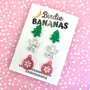 Peut inclure: Un ensemble de six boucles d'oreilles sur le thème de Noël avec un sapin de Noël vert pailleté, un ange argenté pailleté et une boule rose pailletée avec un flocon de neige. Les boucles d'oreilles sont présentées sur une carte blanche avec le texte "Birdie Bananas" et un graphique de banane.