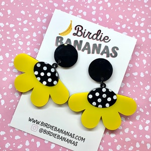 May include: A pair of yellow flower-shaped earrings with black polka dot accents. The earrings are attached to black round discs with a silver post.