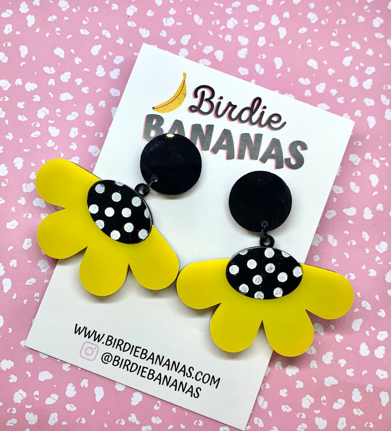 May include: A pair of yellow flower-shaped earrings with black polka dot accents. The earrings are attached to black round discs with a silver post.