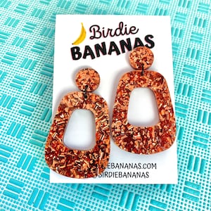 以下が含まれることがあります： 幾何学模様の銅色のグリッターイヤリング。イヤリングは、「Birdie Bananas」の文字とバナナのイラストが描かれた白いカードに載っています。背景は、水色のテクスチャ加工された表面です。