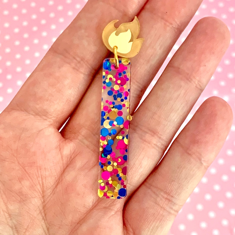May include: A pair of gold earrings with a clear resin bar filled with colorful confetti and a gold flame charm.