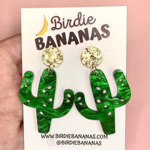 May include: Green cactus-shaped earrings with gold glitter accents. The earrings are on a white card with the text "Birdie BANANAS" and a website address.