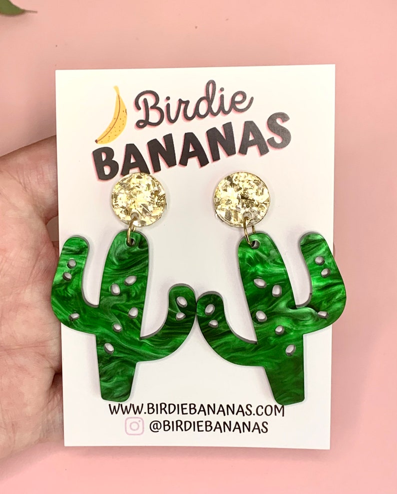 May include: Green cactus-shaped earrings with gold glitter accents. The earrings are on a white card with the text "Birdie BANANAS" and a website address.