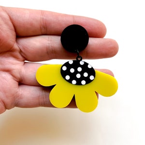 May include: Yellow flower-shaped dangle earrings with a black and white polka dot center. The earrings are made of acrylic and have a black circle at the top.