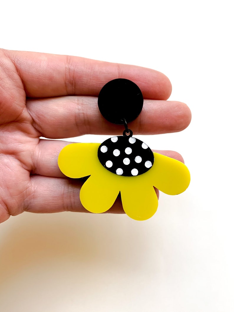 May include: Yellow flower-shaped dangle earrings with a black and white polka dot center. The earrings are made of acrylic and have a black circle at the top.