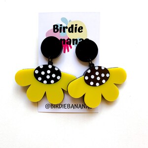 May include: A pair of yellow flower-shaped earrings with black and white polka dot accents. The earrings are hanging from black circular posts. The earrings are on a white card with the text "Birdie Nana" and "@birdiebanana".