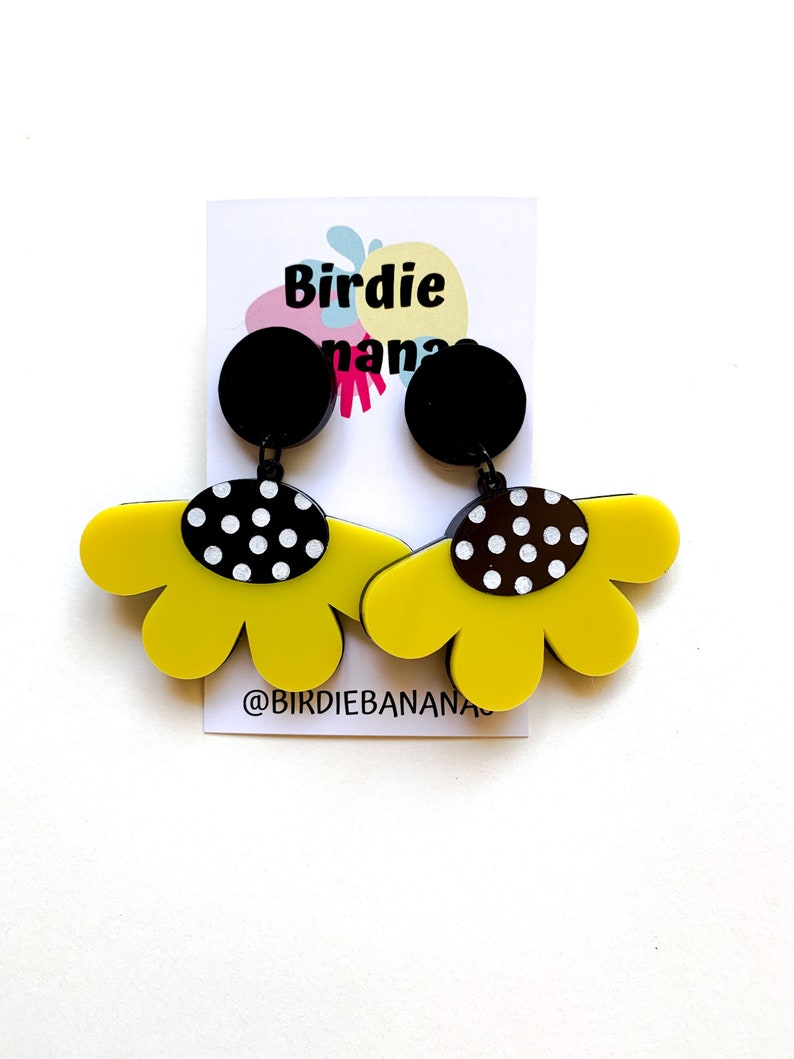 May include: A pair of yellow flower-shaped earrings with black and white polka dot accents. The earrings are hanging from black circular posts. The earrings are on a white card with the text "Birdie Nana" and "@birdiebanana".
