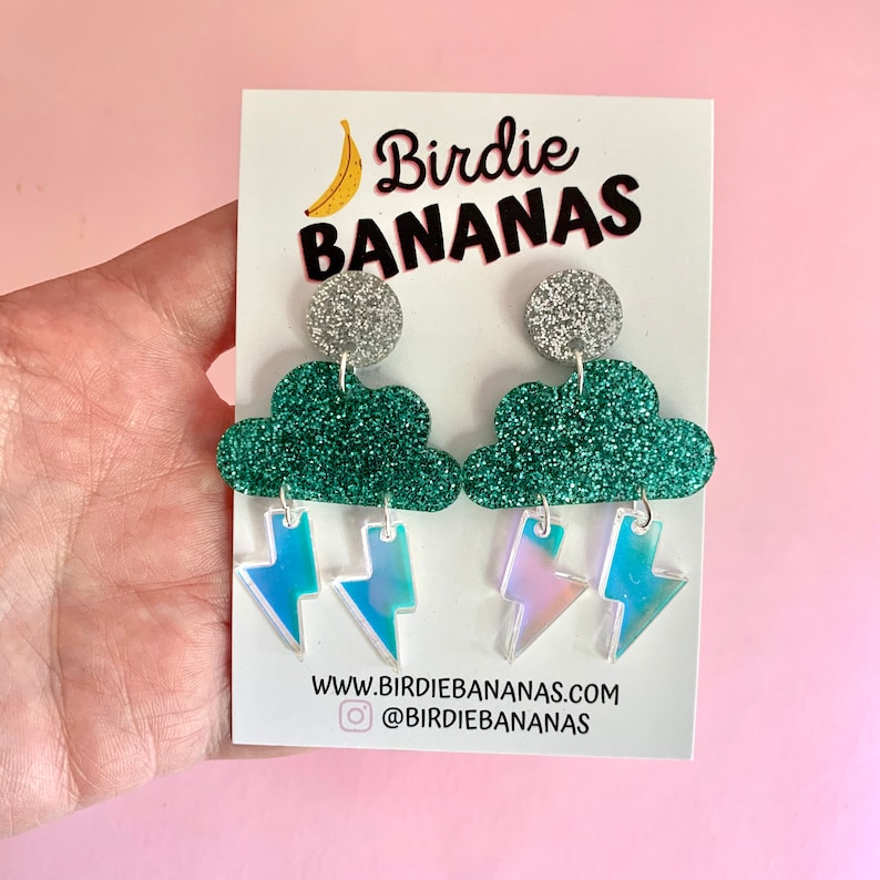 K&ouml;nnte beinhalten: Ein Paar silberne Glitzer-Wolkenohrringe mit regenbogenfarbenen Blitzen als Anh&auml;nger. Die Ohrringe befinden sich auf einer wei&szlig;en Karte mit dem Text "Birdie Bananas" und einer Website-Adresse.