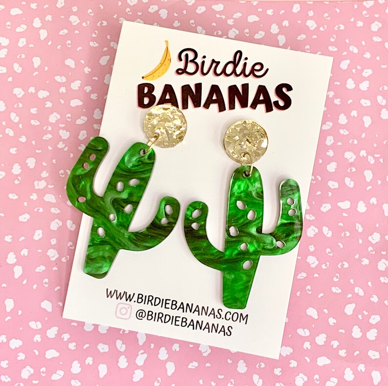 May include: A pair of green cactus-shaped earrings with gold glitter accents. The earrings are on a white card with the text "Birdie BANANAS" and a website address.
