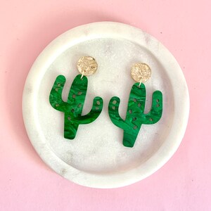 May include: A pair of green cactus-shaped earrings with gold accents. The earrings are made of a marbled green material and have a unique, whimsical design.