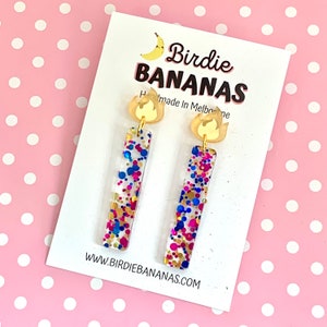 May include: A pair of gold and clear resin earrings with a confetti design. The earrings are shaped like a rectangle and have a gold flame charm at the top. The text "Birdie BANANAS Handmade in Melbourne" is on the card behind the earrings.