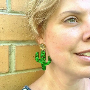 May include: A pair of green cactus-shaped earrings with gold accents. The earrings are made of a textured material and have a unique, whimsical design.