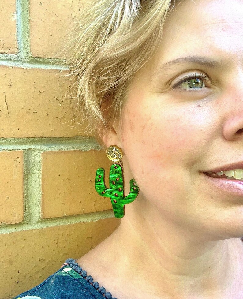 May include: A pair of green cactus-shaped earrings with gold accents. The earrings are made of a textured material and have a unique, whimsical design.