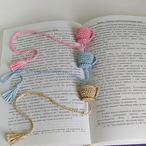 Op de afbeelding: Een open boek met een gehaakt bladwijzer in de vorm van een theekopje. De bladwijzer is lichtblauw en heeft een witte kwast. Het boek is open op een pagina met tekst over boekhouding en bedrijfsleven.