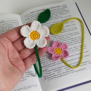 May include: A crocheted bookmark with two flowers and green leaves. The flowers are white with a yellow center and pink with a yellow center. The bookmark is made of yarn and is a great way to mark your place in a book.
