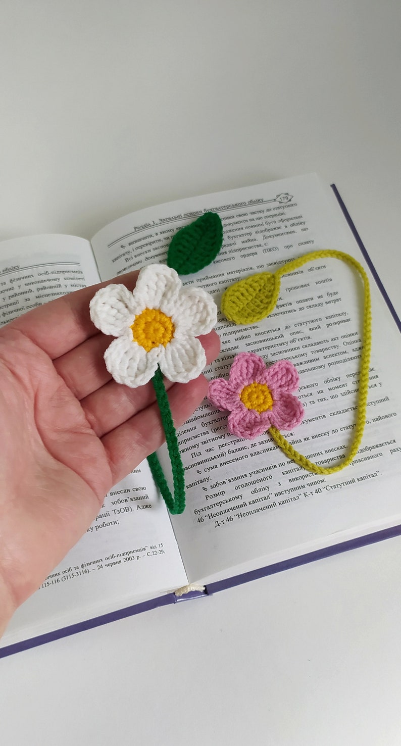 May include: A crocheted bookmark with two flowers and green leaves. The flowers are white with a yellow center and pink with a yellow center. The bookmark is made of yarn and is a great way to mark your place in a book.