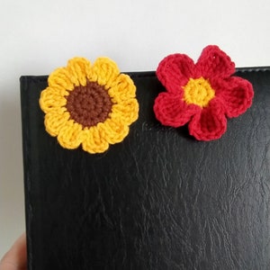 May include: Two crocheted flower embellishments, one yellow with a brown center and one red with a yellow center, on a black surface.