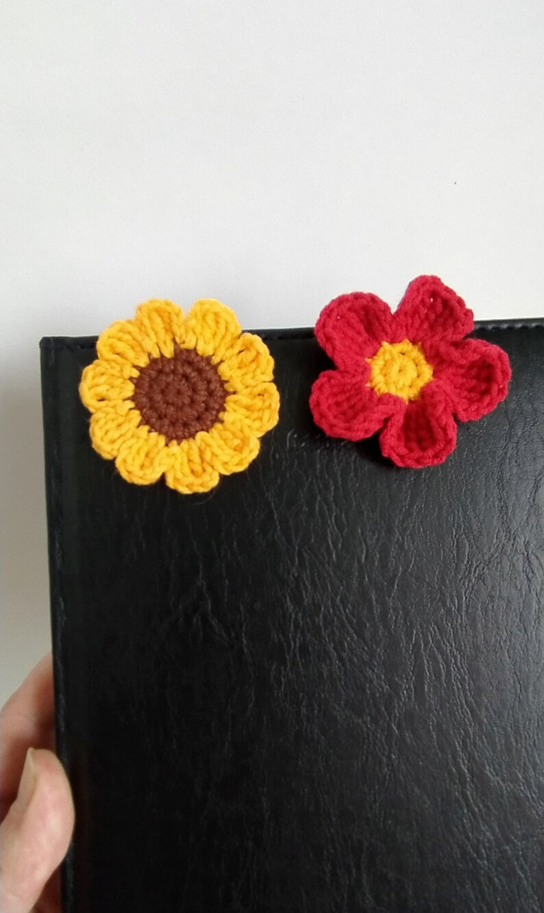 May include: Two crocheted flower embellishments, one yellow with a brown center and one red with a yellow center, on a black surface.
