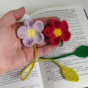 May include: A close-up of a hand holding three crocheted bookmarks. The bookmarks are shaped like flowers with green leaves. The flowers are purple, red, and yellow. The bookmarks are resting on an open book.