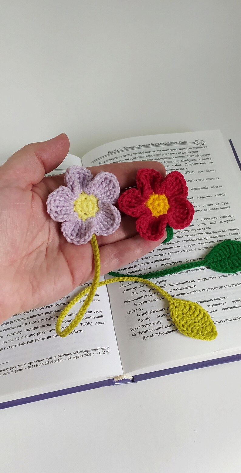 May include: A close-up of a hand holding three crocheted bookmarks. The bookmarks are shaped like flowers with green leaves. The flowers are purple, red, and yellow. The bookmarks are resting on an open book.