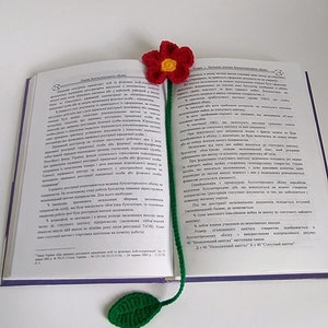 May include: A crocheted bookmark with a red flower and green leaf. The bookmark is placed in an open book with text in a foreign language.