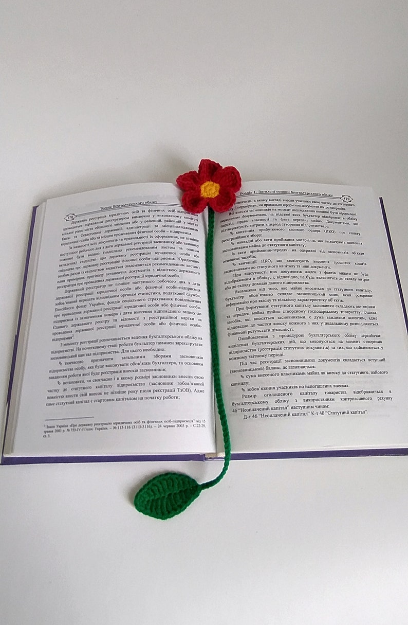 May include: A crocheted bookmark with a red flower and green leaf. The bookmark is placed in an open book with text in a foreign language.