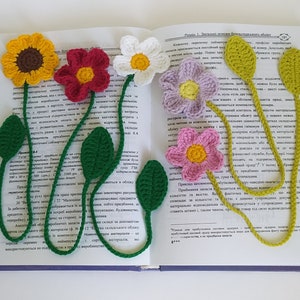 May include: Five crocheted flower bookmarks in different colors, including yellow, red, white, purple, and pink. The bookmarks are attached to a book with the text "Pravila 1. Znachnost' vliyaniya faktorov" visible.