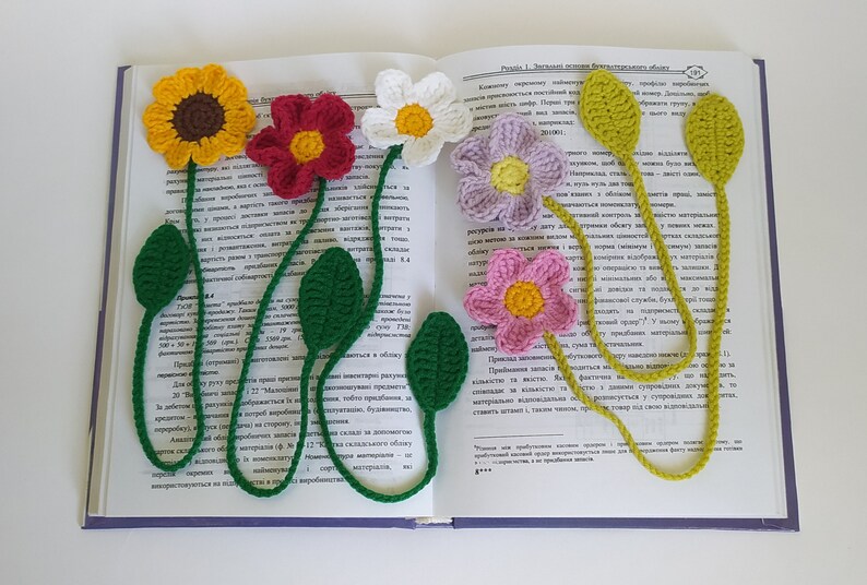 May include: Five crocheted flower bookmarks in different colors, including yellow, red, white, purple, and pink. The bookmarks are attached to a book with the text "Pravila 1. Znachnost' vliyaniya faktorov" visible.