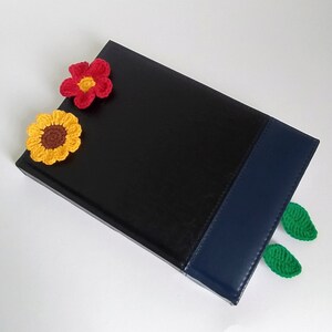 May include: A black leather journal with a blue spine and crocheted flower and leaf embellishments. The flowers are yellow, red, and brown. The leaves are green.