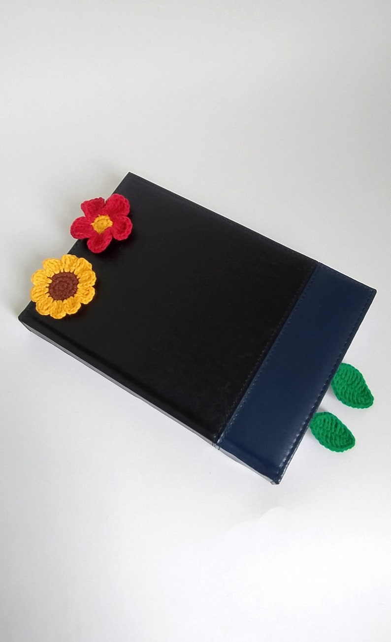 May include: A black leather journal with a blue spine and crocheted flower and leaf embellishments. The flowers are yellow, red, and brown. The leaves are green.